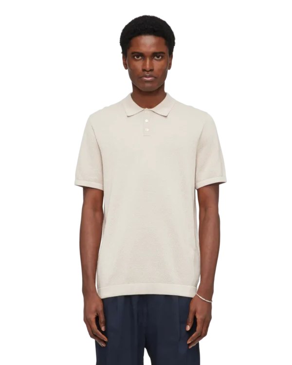 Model draagt DRYKORN polo TRITON in beige, met korte mouwen en fijngebreide structuur.