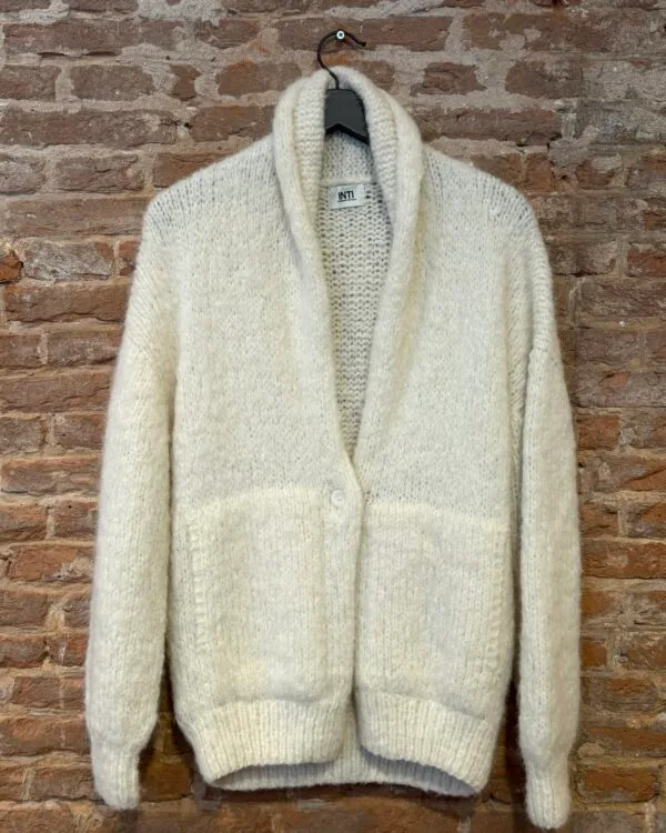 Crèmekleurig handgebreid vest van Inti Knitwear met sjaalkraag en knoopsluiting, gefotografeerd op een hanger tegen een bakstenen muur.