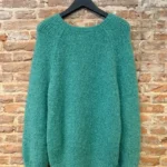 Groene handgebreide trui van Inti Knitwear met ronde hals en lange mouwen, gefotografeerd op een hanger tegen een bakstenen muur.