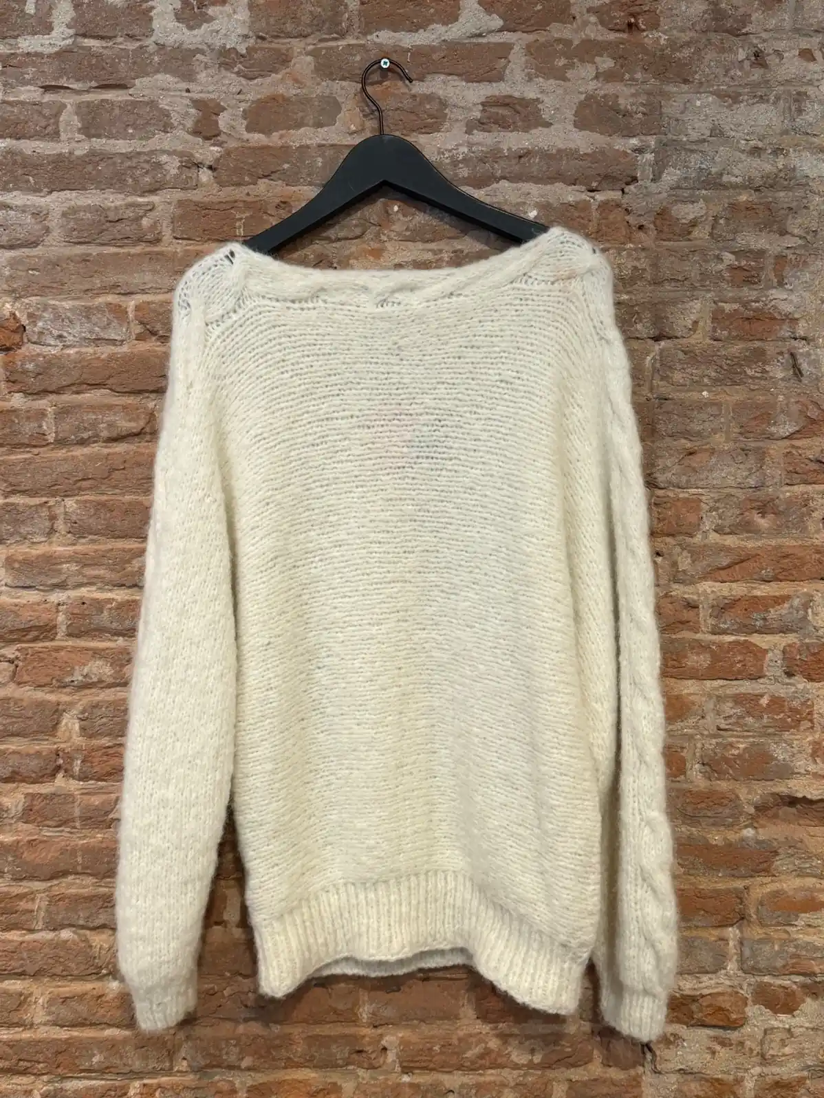 baby-alpaca-wol Witte handgebreide trui van zachte alpaca-wol met ronde hals, gefotografeerd op een hanger tegen een bakstenen muur.